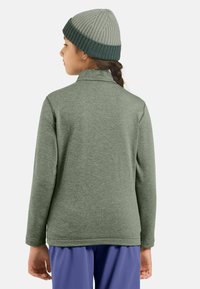 Hellgrünes Langarmoberteil mit Kragen, aus einem strukturierten Stoff. Kombiniert mit einer gerippten, grau-dunkelgrünen Mütze.