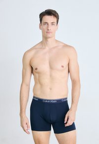 Een man zonder shirt met kort bruin haar draagt donkerblauwe Calvin Klein boxershorts en staat tegen een effen lichte achtergrond.