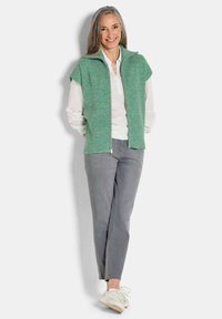 Grüne Strickweste mit Reißverschluss vorne, kurzen Ärmeln und Stehkragen, kombiniert mit einem weißen Hemd und grauen Hosen.