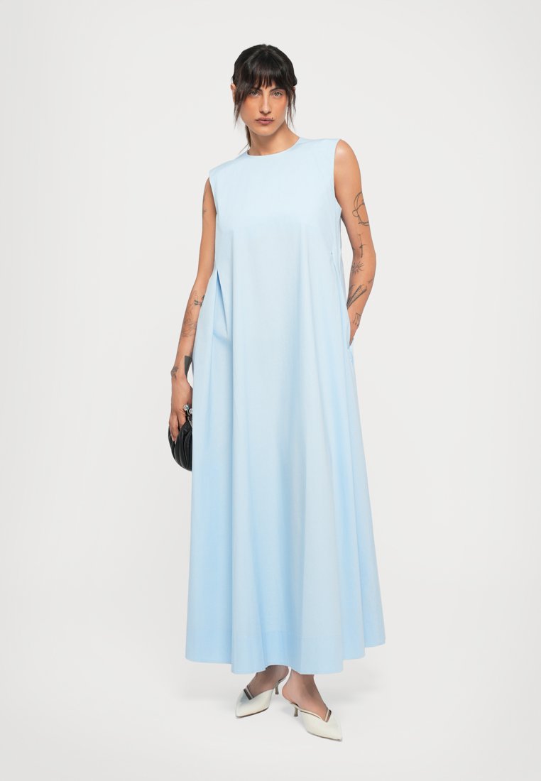 Femme tatouée portant une robe maxi sans manches bleu clair, des escarpins blancs à bout pointu et tenant un petit sac à main noir.