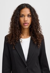 Černý blazer s klopou a jediným knoflíkem, nošený přes bílou košili. Model má dlouhé, kudrnaté vlasy a neutrální výraz.