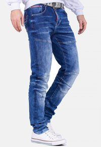 Jean en denim bleu avec une texture abîmée, plusieurs poches et des coutures contrastantes. Comprend une ceinture avec une boucle visible.