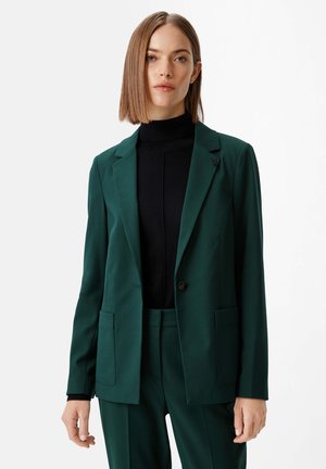 Grüner maßgeschneiderter Blazer aus glattem Stoff mit einem Knopfverschluss, zwei Taschen vorne und einem Reverskragen. Kombiniert mit einem schwarzen Rollkragenpullover.