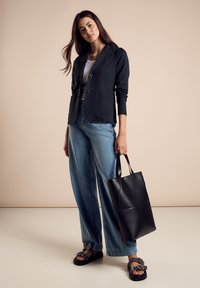 Schwarze Blazer mit zwei vorderen Taschen, getragen über einem grauen Oberteil. Hochtaillierte blaue Jeans, schwarze Tragetasche und dunkle Sandalen mit dekorativen Schnallen.