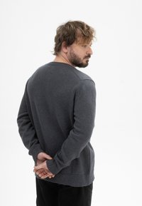Grauer Strickpullover mit klassischem Design, rundem Halsausschnitt, gerippten Bündchen und Saum, über einer dunklen Hose getragen, mit hinter dem Rücken verschränkten Händen.
