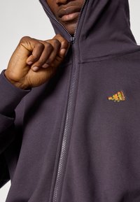 Corta-vento roxo escuro com fecho éclair, bolso canguru, punhos em malha e um pequeno emblema multicolorido no lado esquerdo do peito. Textura suave.