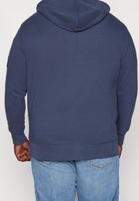Felpa blu marina con cappuccio regolabile, polsini e orlo a costine. Presenta linee pulite e una finitura testurizzata. Vista posteriore senza loghi o motivi.