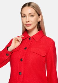 Rode wolmix jas met een kraag, knopen aan de voorkant, twee borstzakken en lange mouwen. Heeft een gestructureerde afwerking en zwarte knoopaccenten.