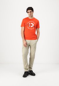 Orange bomuld t-shirt med trykt tekst og logo, parret med beige cargo bukser. Sorte sko fuldender outfittet.