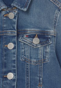 Denimová bunda ve středně modré barvě s kovovými knoflíky, dvěma náprsními kapsami a kontrastním žlutým prošíváním podél švů.