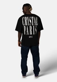 Černé volné tričko s bílým nápisem "CRYSTAL PARIS" a "DEPT.75" na zadní straně, ve spojení s modrými džíny a fialovými teniskami.