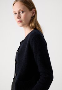 Frau mit heller Haut und glattem, hellbraunem Haar trägt eine schwarze, strukturierte, geknöpfte Strickjacke vor einem schlichten weißen Hintergrund.