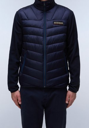 Mężczyzna w granatowej pikowanej kamizelce bez rękawów nałożonej na długi rękaw, stojący z rękami wzdłuż ciała na jednolitym tle.