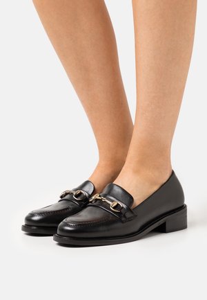 Mocassins en cuir noir avec un talon bas, présentant une accent en métal doré et des coutures détaillées sur la tige. Texture lisse.