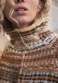 Pull en tricot avec col montant, dans des tons chauds de marron, crème et bleu clair, avec un design texturé et à motifs, ainsi que des détails de coutures apparentes.