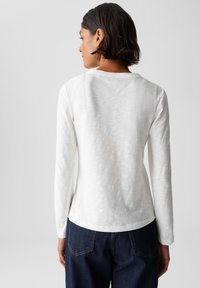 Donna con i capelli corti e scuri, che indossa una semplice camicia bianca a maniche lunghe e jeans blu scuro, vista di spalle su uno sfondo uniforme.