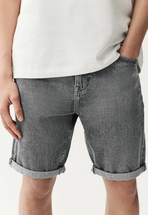 Shorts en denim gris avec des revers, présentant un design classique à cinq poches et une texture subtile, associés à une chemise blanche.