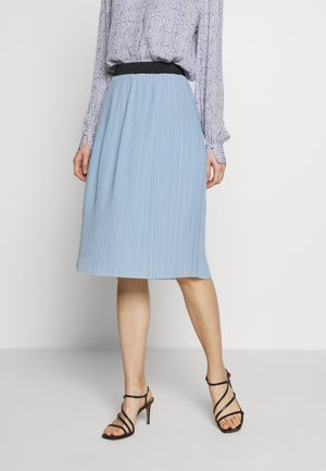 Femme portant une jupe midi plissée bleu clair, un chemisier à manches longues à motifs et des sandales à talons hauts strappées noires, se tenant devant un fond uniforme.