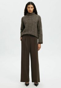 Femme portant un pull marron à motif chevrons avec un col montant, un pantalon marron foncé à jambes larges et des chaussures noires pointues, debout sur fond blanc.