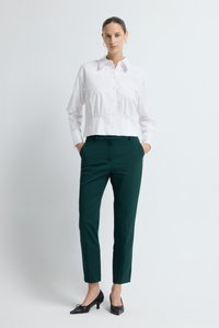 Camicia bianca corta con bottoni e colletto, abbinata a pantaloni sartoriali verde scuro e scarpe nere con tacco kitten. Tessuto dalla texture liscia.