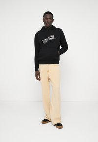 Svart hoodie med grafiskt tryck, beiga vida jeans och svarta sneakers med bruna sulor, stående mot en neutral vit bakgrund.