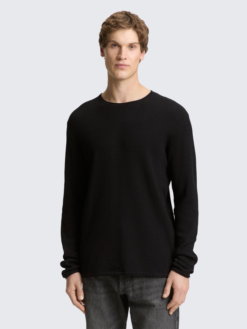 Fekete, hosszú ujjú bordázott kötött pulóver kerek nyakkal és kényelmes szabással, sötét farmernadrággal párosítva, világos háttér előtt bemutatva.