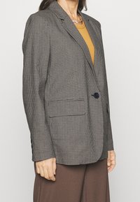 Blazer à carreaux noir et blanc, avec une fermeture à un bouton, des revers crantés et deux poches avant. Associé à un haut couleur moutarde.