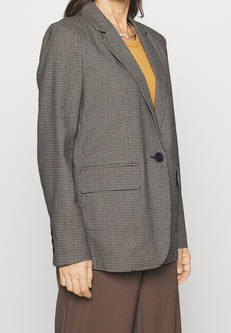 Blazer à carreaux noir et blanc, avec une fermeture à un bouton, des revers crantés et deux poches avant. Associé à un haut couleur moutarde.