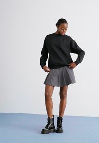 Sudadera negra con cuello redondo, combinada con una falda de tablas gris. La modelo lleva botas negras de cordones gruesos. Textura suave en general.