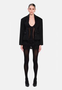 Musta räätälöity takki, jossa on ylisuuria kauluskäänteitä, läpikuultava musta paita, röyhelöinen musta minihame, läpikuultavat sukkahousut ja mustat piikkikorkokengät.