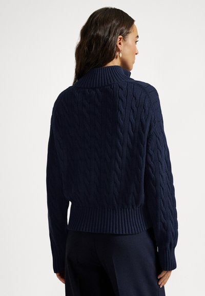 Γυναίκα που φοράει μπλε σκούρο πουλόβερ με ψηλό γιακά και σχέδιο πλεξούδας, καθώς και σκούρο παντελόνι, εμφανιζόμενη από πίσω με φόντο λευκό.