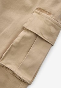 Beige bomullstyg med en strukturerad yta, med en rullad manschett och kontrasterande sömmar. Diskret varumärkning är synlig.