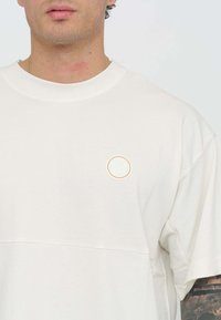 T-shirt in cotone color avorio con una patch a logo rotondo in colori delicati, maniche corte e una vestibilità rilassata con un dettaglio di cucitura orizzontale.