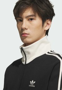 Crna zip-up jakna s bijelim ovratnikom i prugama, teksturirana tkanina, s malim srebrnim logotipom na prsima.