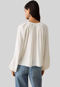 Leichte weiße Bluse mit gerafftem Ausschnitt und langen Ärmeln, kombiniert mit blauen Jeans, zeigt eine lockere Passform und weiche Textur.