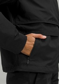 Crna jakna izrađena od mekanog, laganog materijala, sa patent-zatvaračem na bočnoj strani i opuštenim krojem s blagim sjajem i teksturiranim detaljima šivanja.