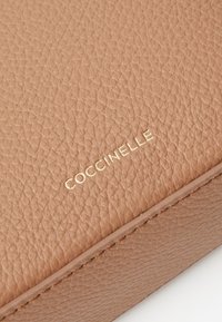 Tumši ādas maks ar strukturētu virsmu. Izceļas ar zelta iespiešanu "COCCINELLE" apakšējā stūrī, ar redzamām šuvēm.