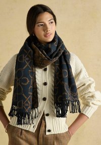 Tmavomodrý šál s hnedým geometrickým vzorom a strapcami, nosený cez krémový pletený kardigán s veľkými gombíkmi a textúrovanými rukávmi.