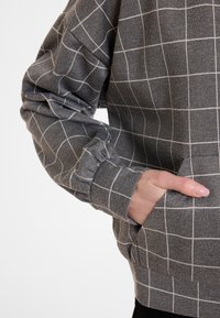 Sweat-shirt gris avec un motif à carreaux blancs, présentant des manches côtelées et une poche avant. Fabriqué en tissu doux et texturé.
