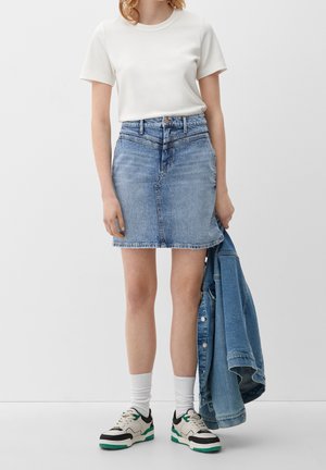 Kobieta w białym t-shircie, niebieskiej dżinsowej spódnicy, białych skarpetkach i trampkach, trzymająca niebieską dżinsową kurtkę przy boku.