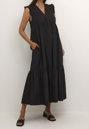 Vrouw draagt een zwarte mouwloze maxi-jurk met ruches aan de schouders, een V-hals, zakken en zwarte open teen sandalen met blokhak.