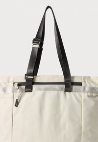 Sac fourre-tout beige avec une surface texturée, des sangles noires réglables, une fermeture éclair sur le dessus et une bande d'accent argentée. Il dispose d'une poche zippée sur le côté.