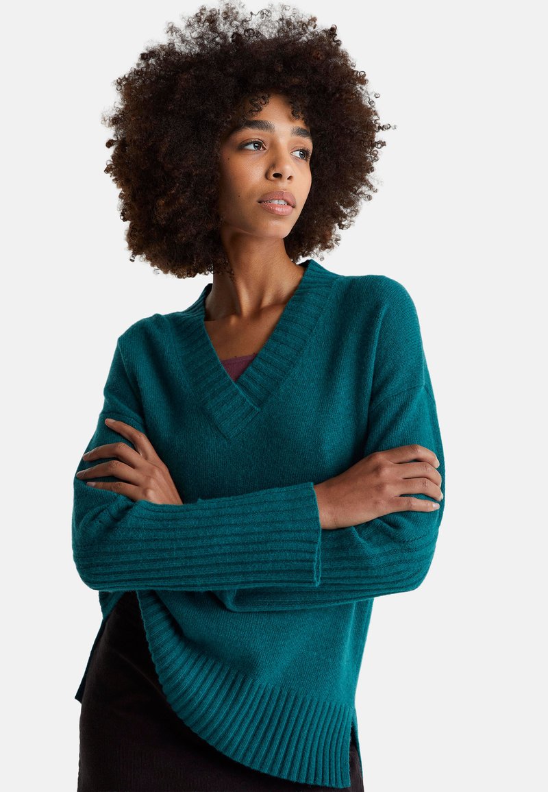 Maglione teal a scollo a V realizzato in materiale morbido, con dettagli a coste sulle maniche e sull'orlo, vestibilità comoda e spacchi laterali.