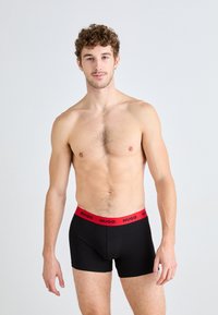 Black boxer briefs with a red elastic waistband labeled "HUGO." The material appears smooth, fitted, and stretching slightly for comfort.