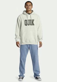 Svetilka svetlo siva s kapuco z napisom "QUIK" v debelih črnih črkah, v kombinaciji z ohlapnimi svetlo modrimi kordnimi hlačami in črnimi športnimi copati.