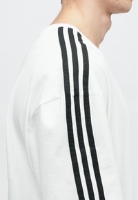 White long-sleeve shirt with black three-stripe design on the shoulder, featuring a relaxed fit and soft cotton material.