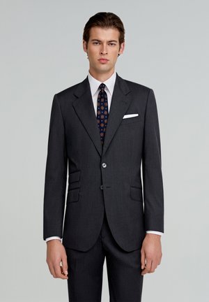 Grau maßgeschneiderter Anzug mit zwei vorderen Taschen, einem Knopfverschluss und Reverskragen. Weißes Hemd und gemusterte Krawatte ergänzen das Outfit.