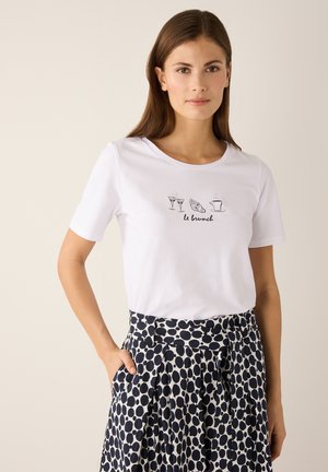 Junge Frau trägt ein weißes T-Shirt mit Brunch-Motiven und einen schwarz-weiß gepunkteten Rock, posiert mit einer Hand in der Tasche.