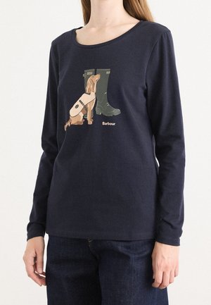 Kobieta w granatowej koszuli z długim rękawem z grafiką przedstawiającą siedzącego brązowego psa obok wysokich zielonych kaloszy oraz napisem „Barbour” na przodzie.