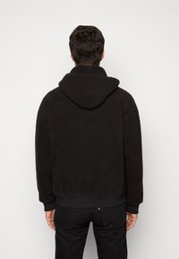 Mann in schwarzem Kapuzenfleecejacke und schwarzen Jeans, mit dem Rücken zur Kamera vor einem schlichten weißen Hintergrund stehend.